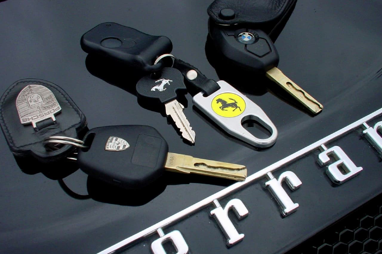 Collection of vintage metal car keys from luxury brands including Porsche, BMW, and Ferrari displayed on dark surface