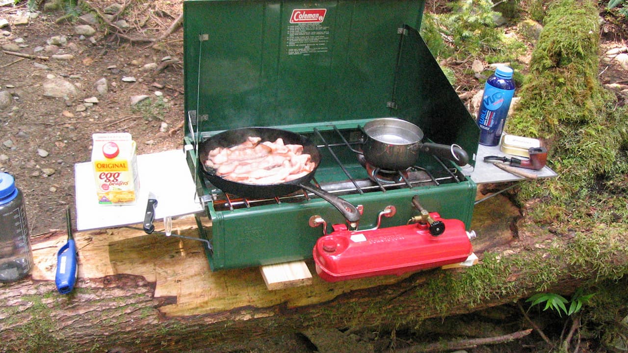 Green Coleman portable camping stove with bacon cooking in pan, red fuel tank, and camping supplies on wooden table