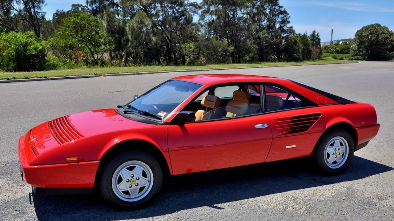 Red Ferrari Mondial coupe with distinctive side strakes and tan interior parked on asphalt road with trees