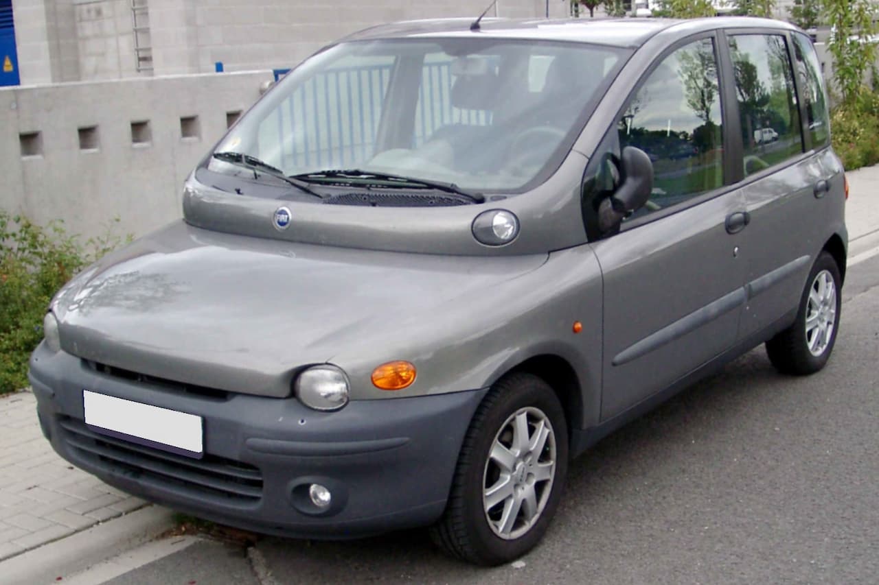 Gray Fiat Multipla minivan parked on street with distinctive wide windshield design, alloy wheels, and concrete wall behind