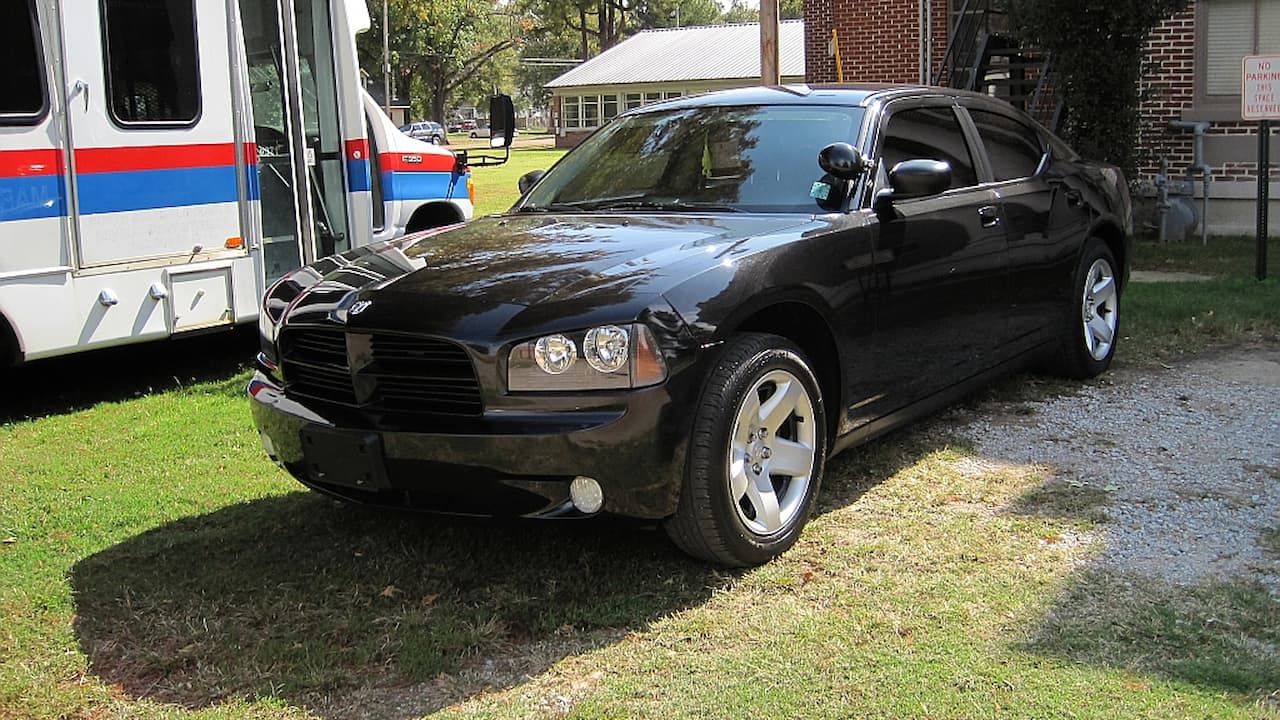 Black Dodge Charger with heavily tinted windows parked on grass near ambulance and brick building in daylight
