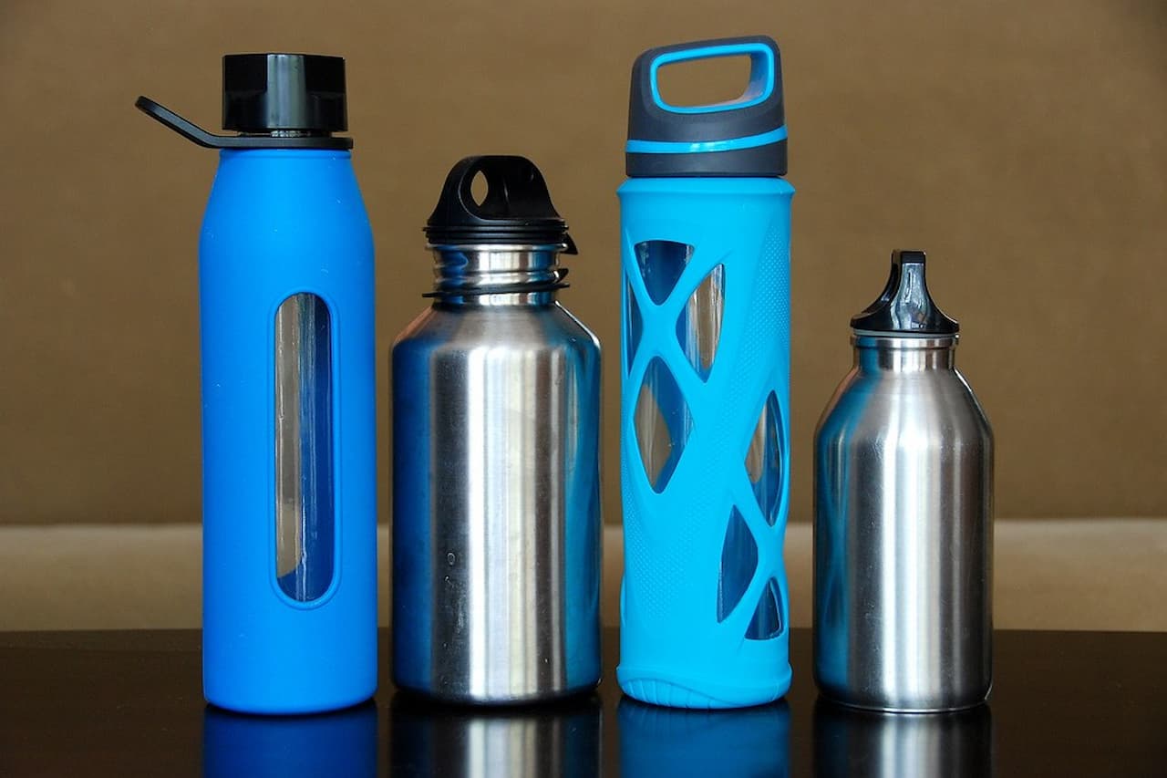 Four reusable water bottles of different styles and colors arranged on a reflective surface against a beige background