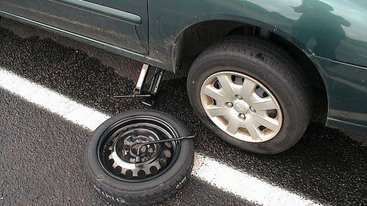 Car jack and spare tire on asphalt road next to dark green vehicle during roadside tire change
