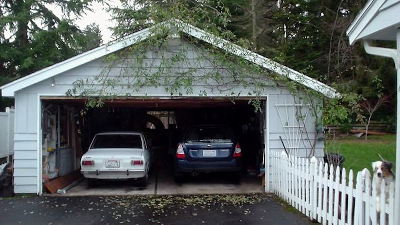 Two-car garage with open door showing white and blue cars parked inside, white picket fence in foreground