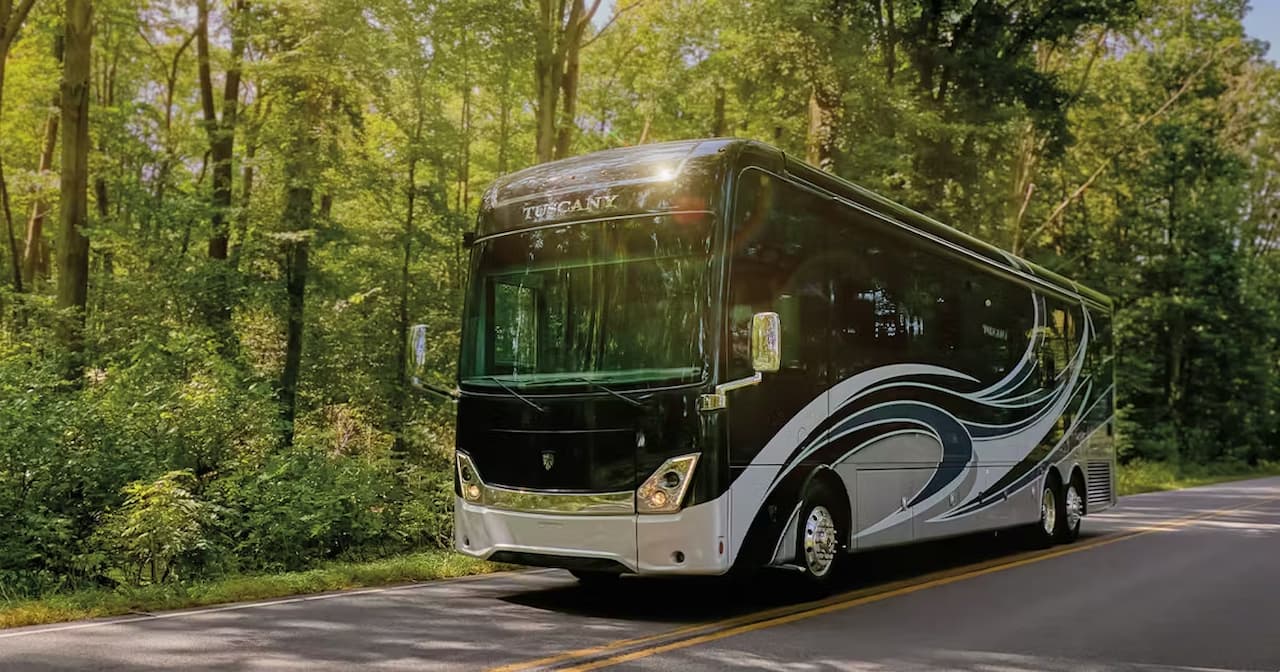 Black and gray Thor Motor Coach Tuscany luxury RV driving on tree-lined asphalt road through forested area