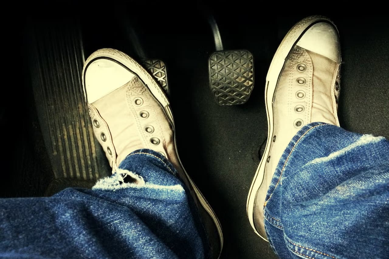 Driver's feet wearing white sneakers and blue jeans positioned on car pedals, showing gas and clutch pedal operation