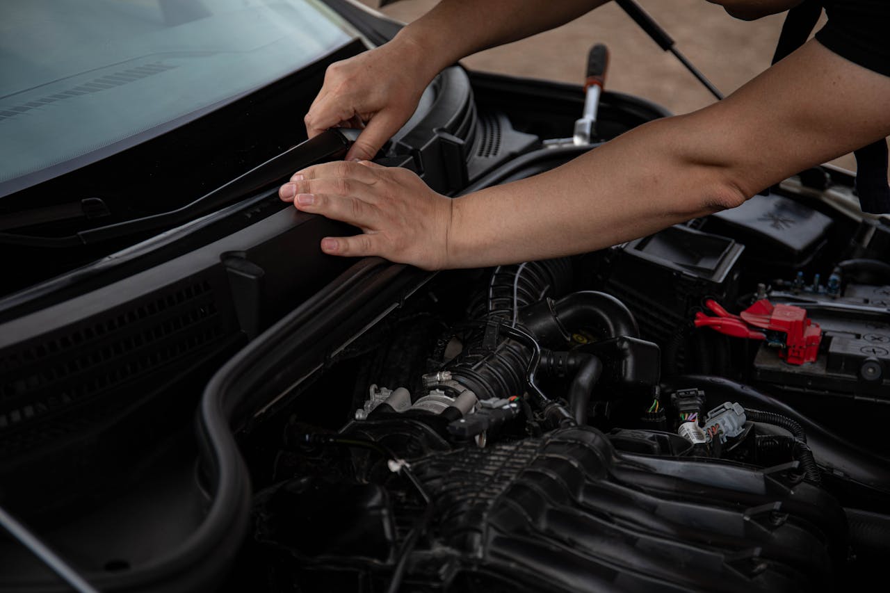 Person's hands working under open car hood, accessing engine compartment with visible wiper motor and mechanical components
