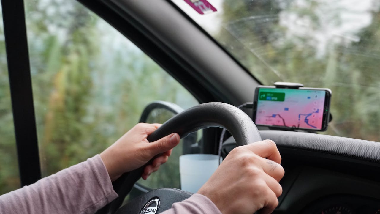 Driver's hands on steering wheel in car with smartphone mounted on dashboard displaying navigation app, trees visible outside