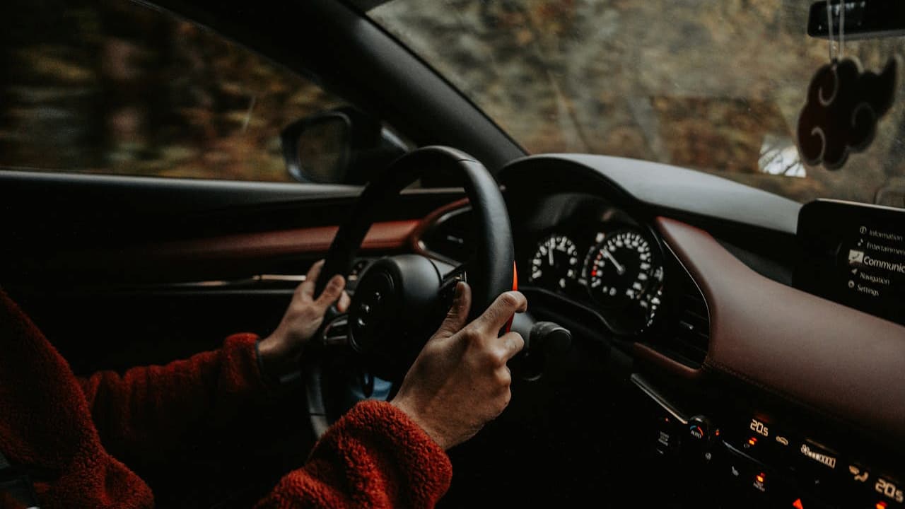 Driver in red sweater gripping steering wheel inside car with analog gauges and infotainment screen visible