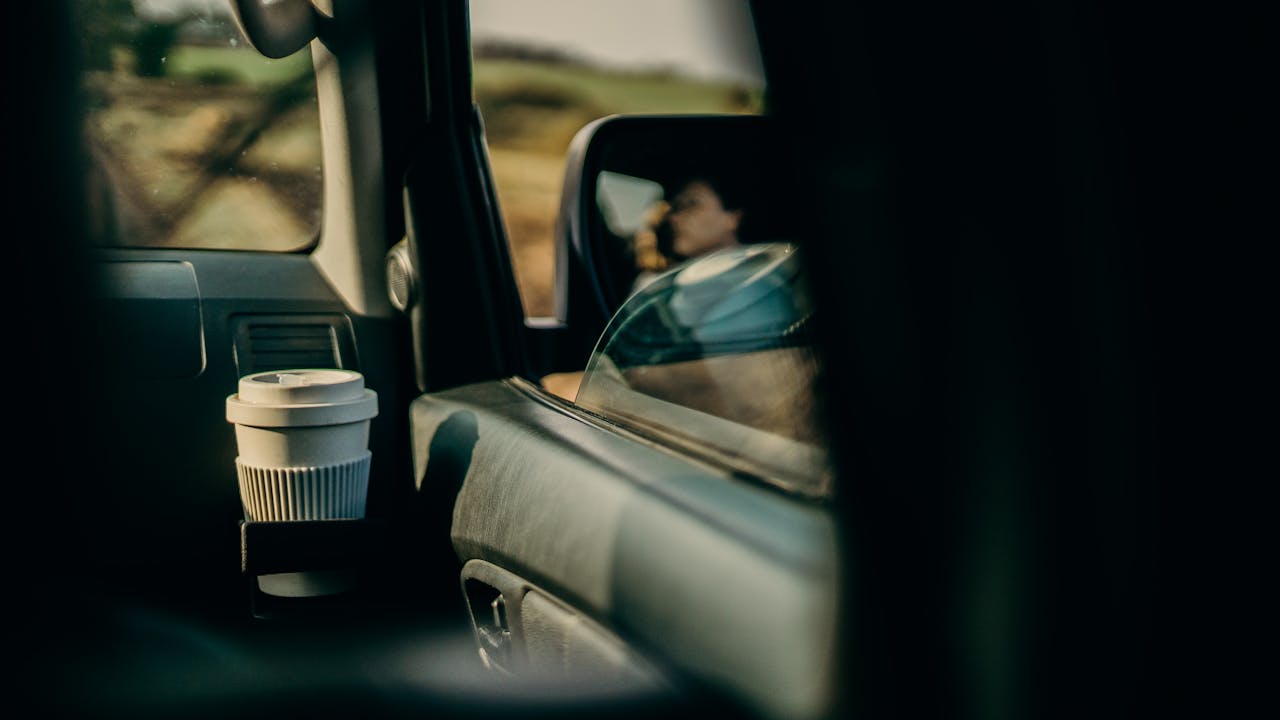 Car interior showing coffee cup in center console cup holder, with driver visible in rearview mirror during daylight