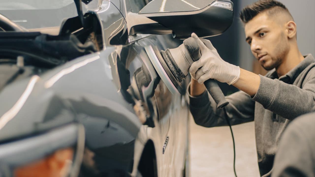 Man in gray shirt and white gloves using circular polishing tool on silver car's surface in garage workshop