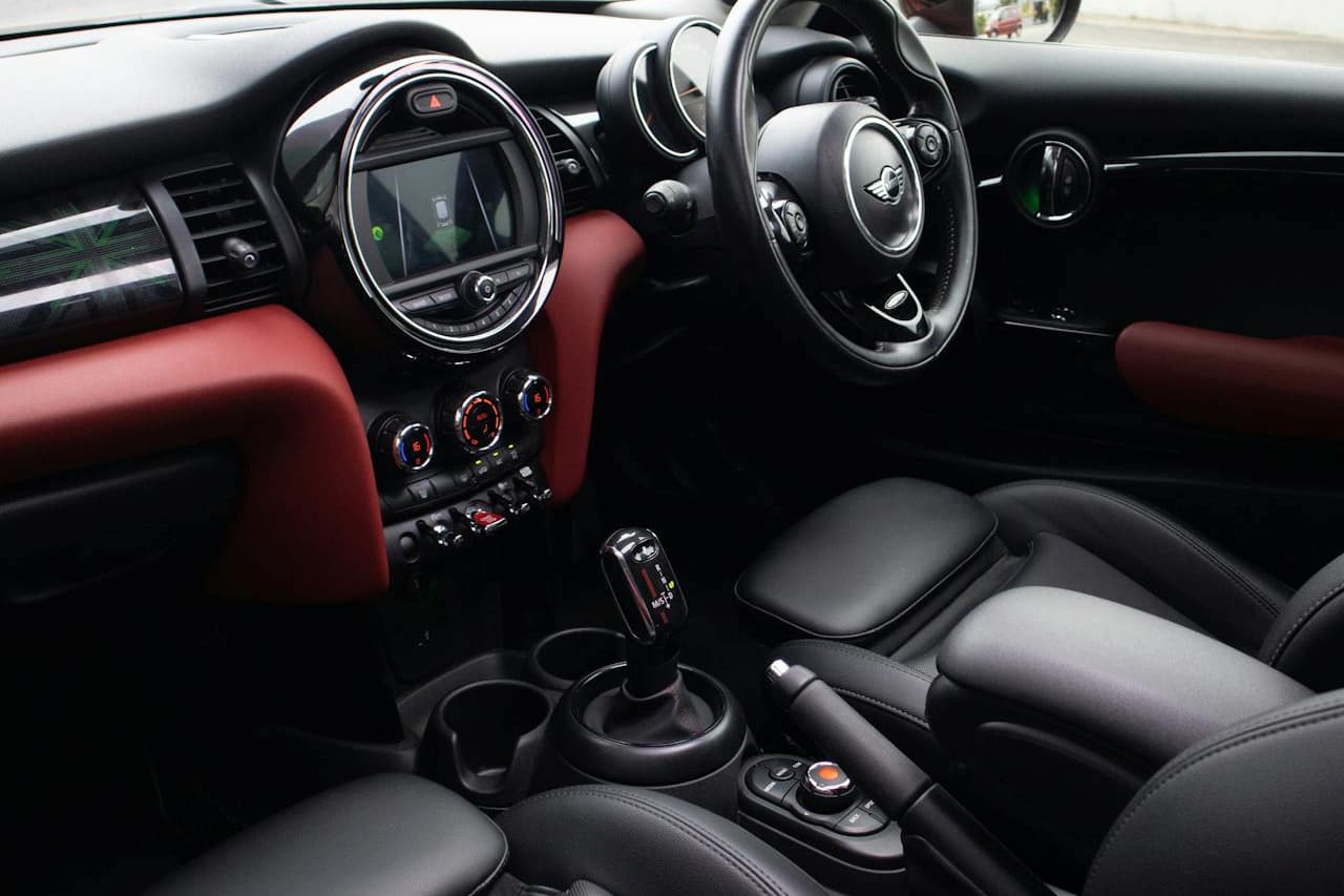 Mini Cooper interior showing dashboard with circular display, steering wheel, red door panels, black leather seats, and manual gear shifter