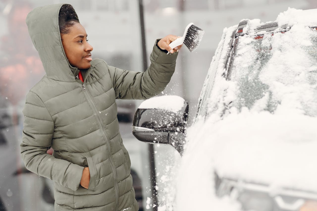 A woman in olive green winter jacket uses an ice scraper to clear snow from her car windshield