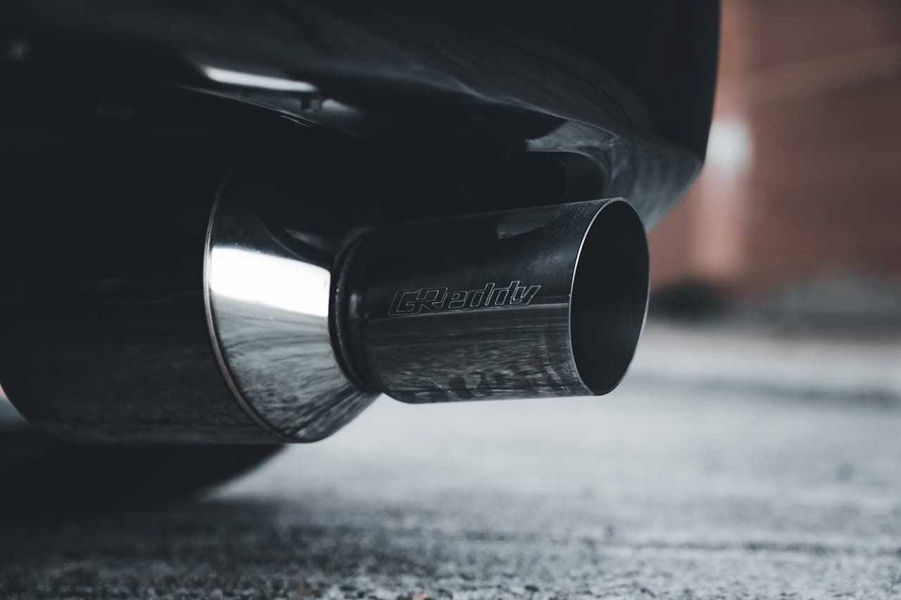 Close-up of black GReddy exhaust tip with carbon fiber finish, positioned low above asphalt pavement surface