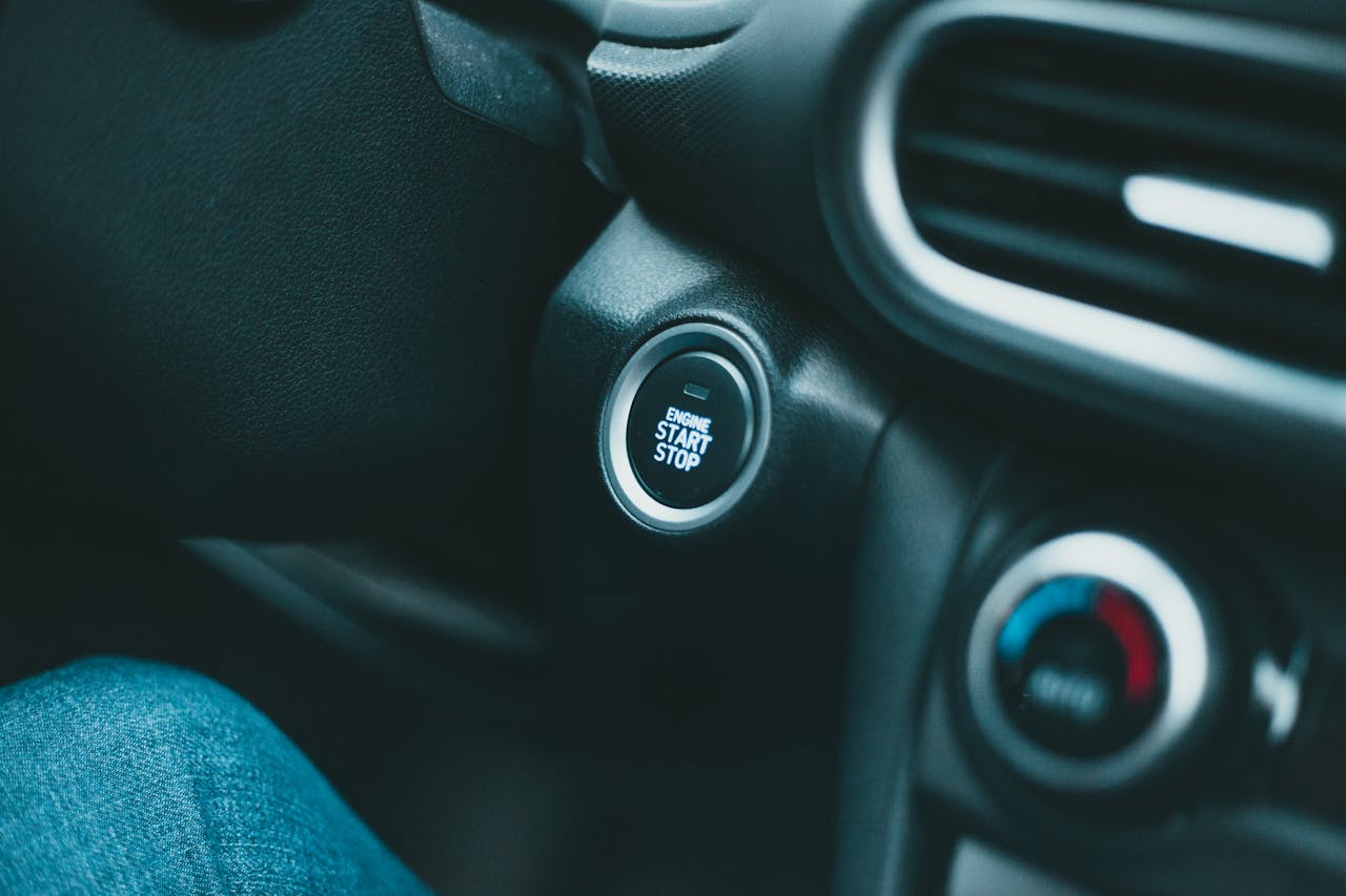 Car dashboard showing illuminated "ENGINE START STOP" button with blue light, surrounded by climate control knobs and air vents