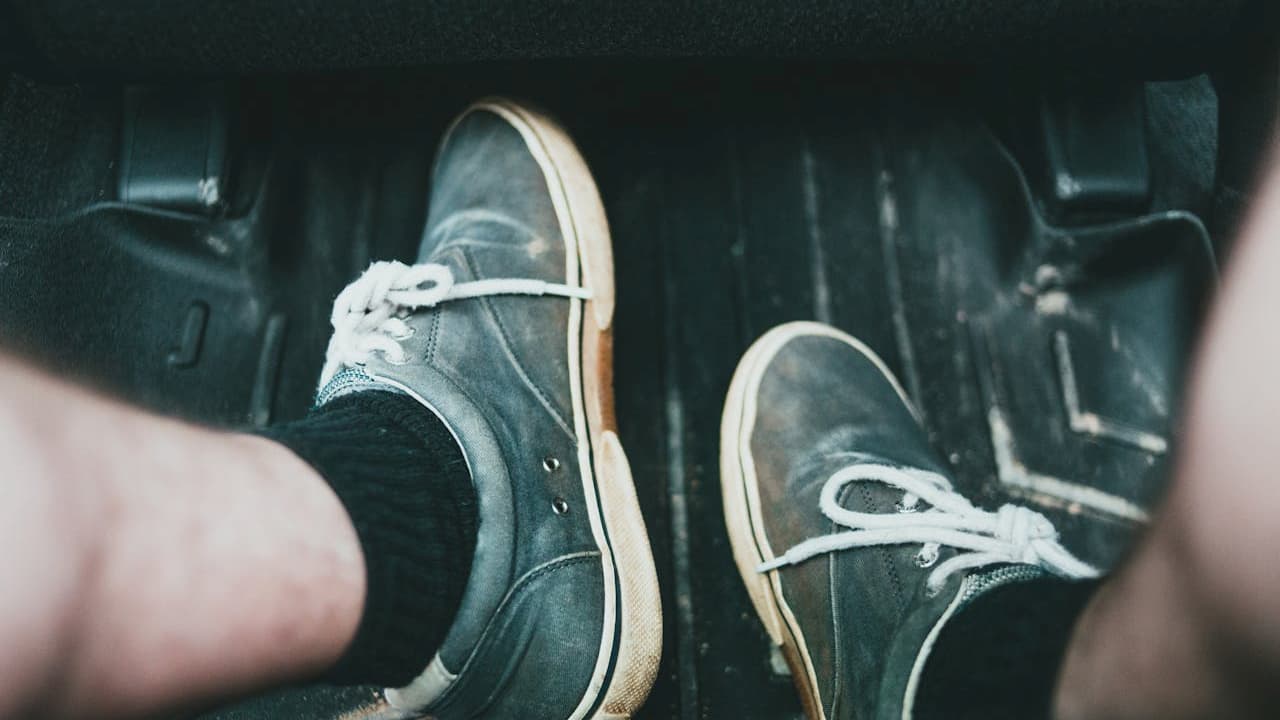 Person's feet wearing gray canvas sneakers with white laces positioned on black rubber car floor mats inside vehicle