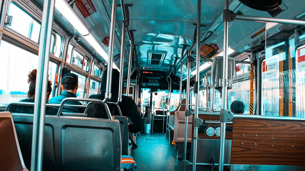 Interior of a city bus with passengers seated, metal handrails, large windows, and blue-tinted lighting throughout the vehicle