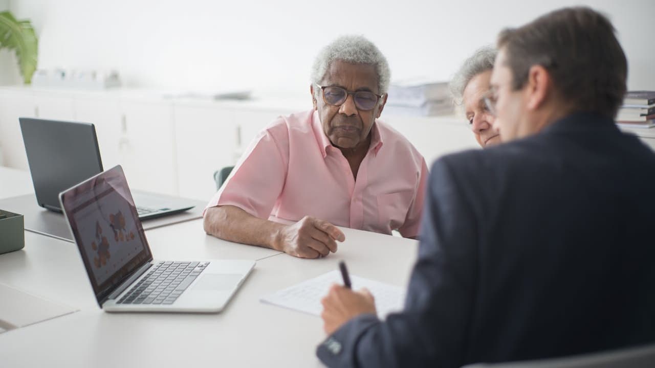 Elderly man in pink shirt and glasses sits at desk with laptop, meeting with younger man in dark suit