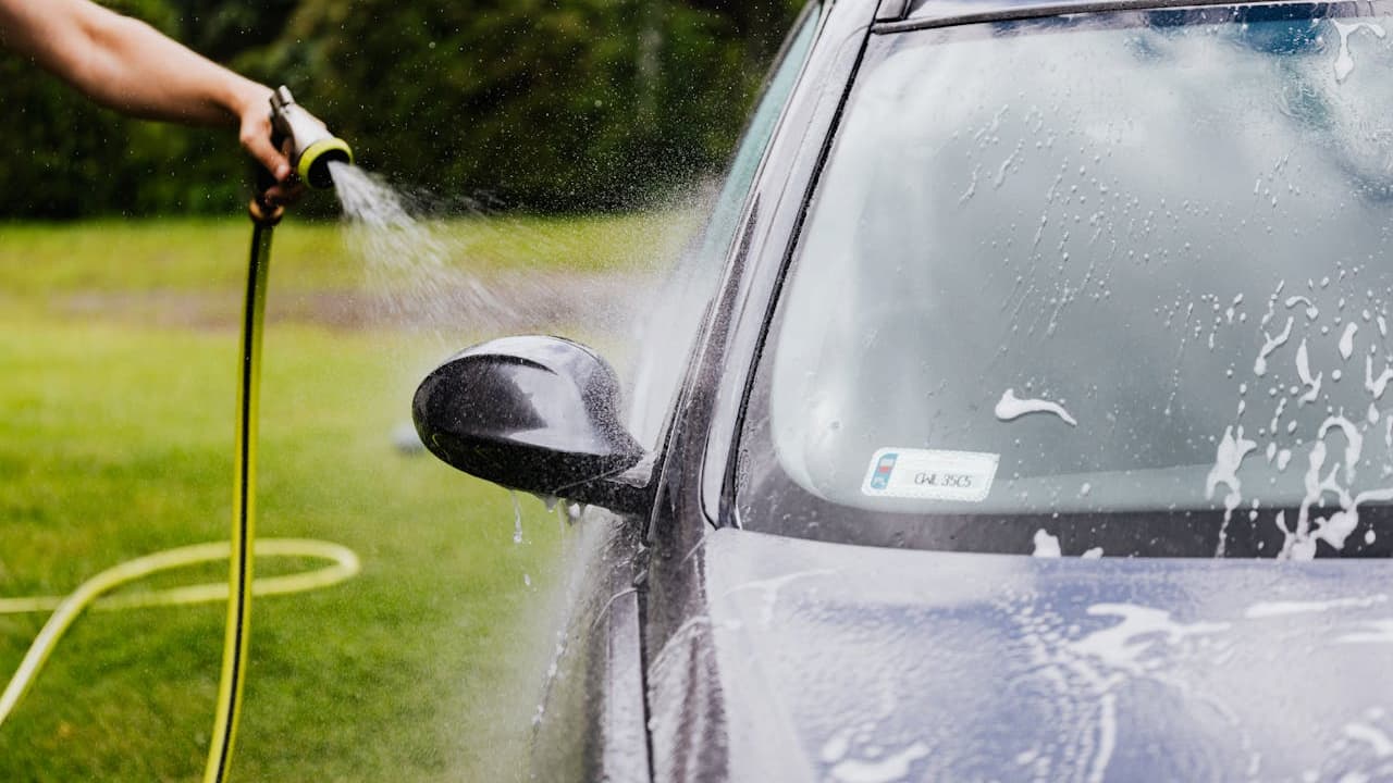 Person washing gray car with pressure washer hose, water spray visible on vehicle windows and side mirror