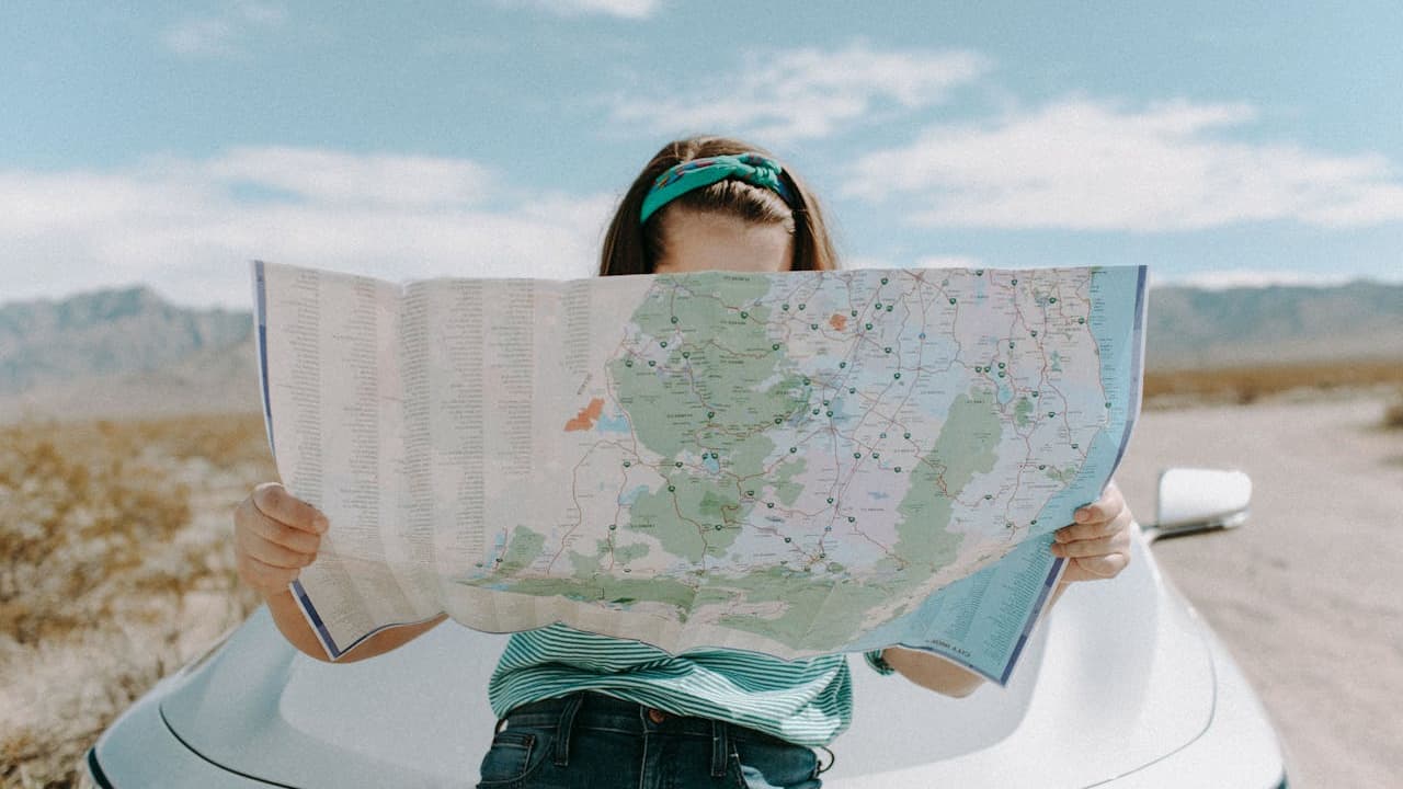 A woman with green headband holds an open road map while sitting on white car in desert landscape