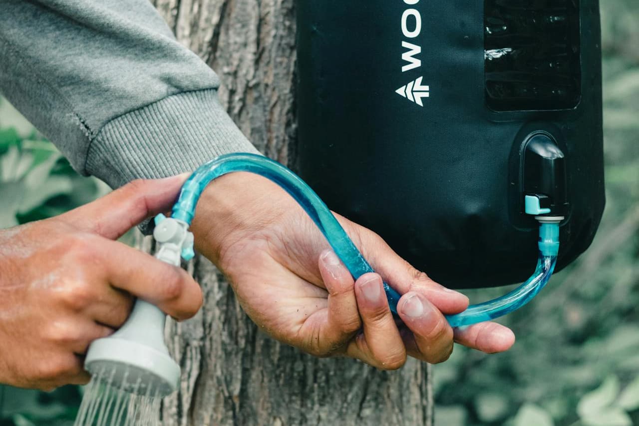 Person holding blue curved water tube from black portable shower device against tree trunk in outdoor setting