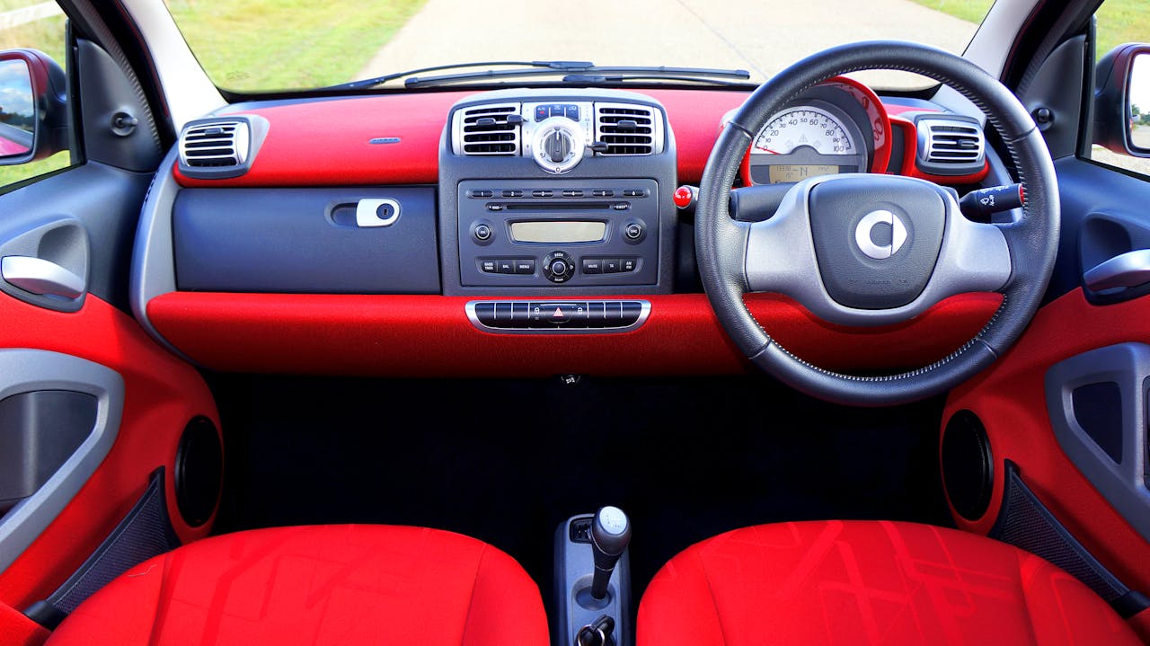 Car interior with red and gray dashboard, steering wheel, manual transmission, radio controls, and safety instrumentation visible