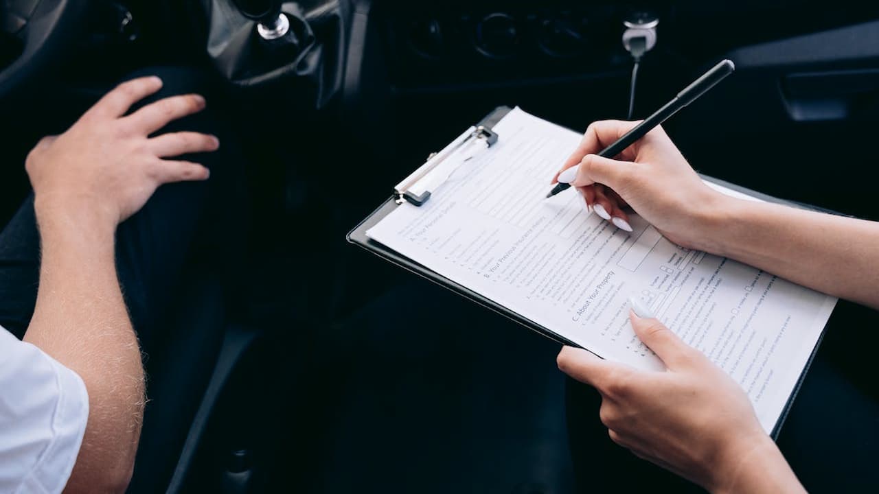 Person in car interior writing on clipboard with insurance forms while gesturing with other hand