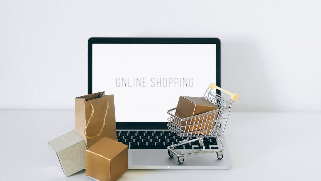 Laptop displaying "Online Shopping" with miniature shopping cart, cardboard boxes, and shopping bag on white background