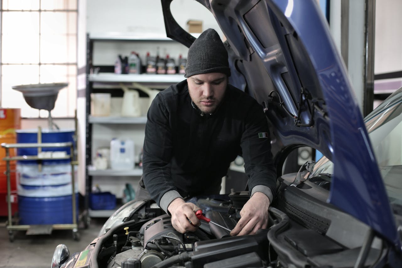 A man in black beanie and shirt working on blue car engine with screwdriver in professional garage workshop