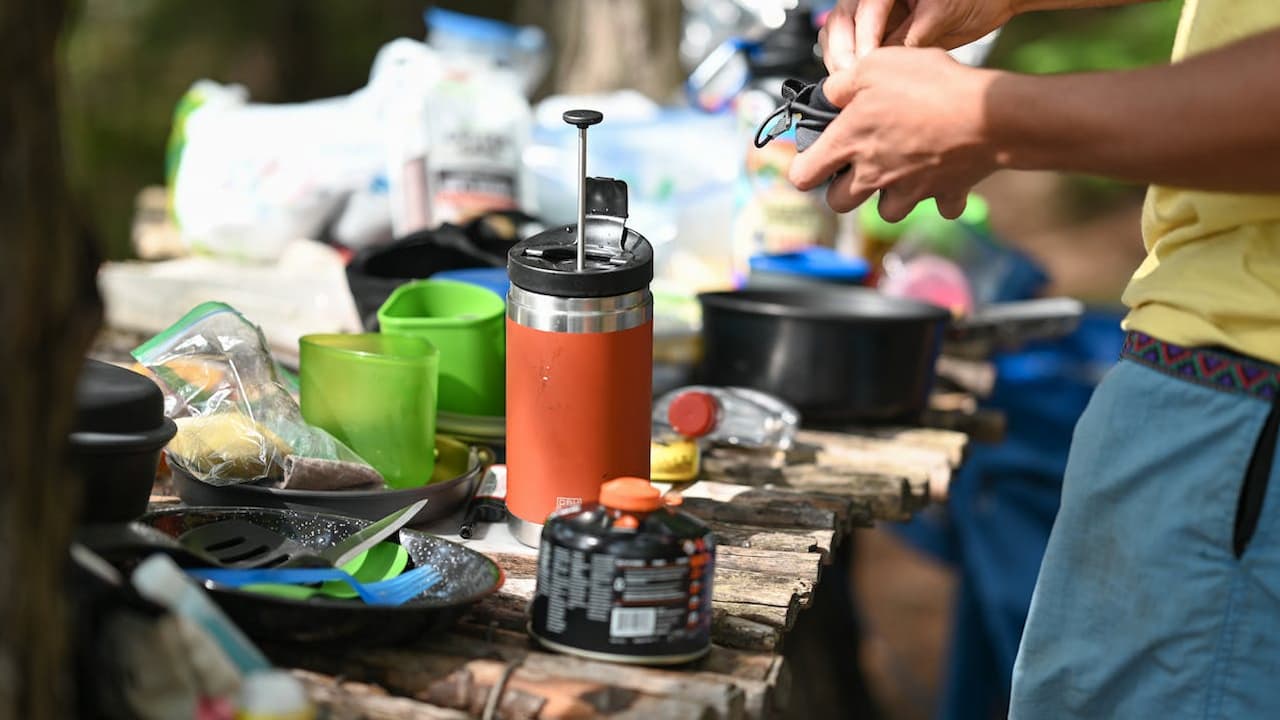 Person using orange camping coffee press with green cups, cookware, and camping gear scattered on wooden outdoor table