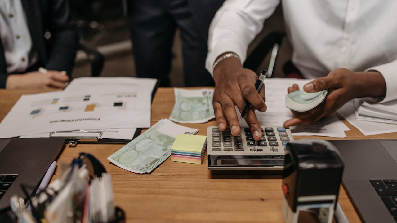 Person using calculator at wooden desk with cash, documents, and office supplies during financial planning session