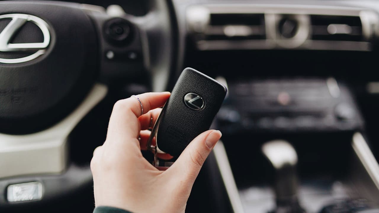 Hand holding black Lexus key fob with textured surface and brand logo, with blurred car interior dashboard background