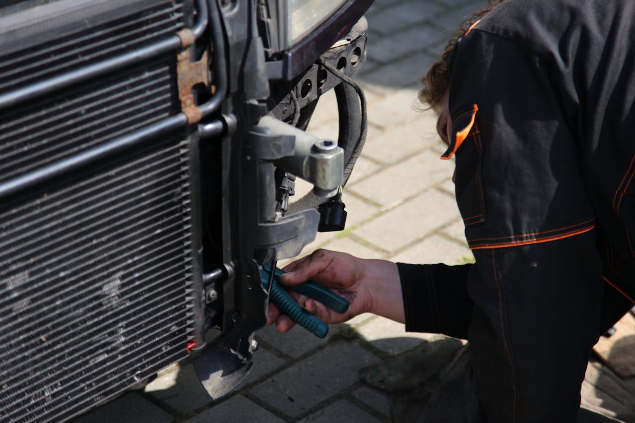 Mechanic working on car radiator with cooling fins, holding green tool while performing maintenance in automotive repair shop