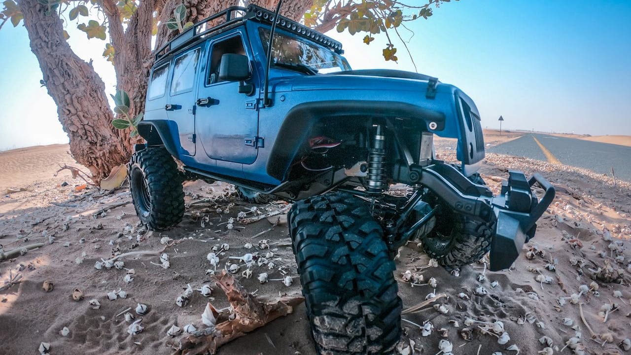 Blue off-road vehicle with exposed suspension system parked under desert tree, showing large tires and mechanical components underneath