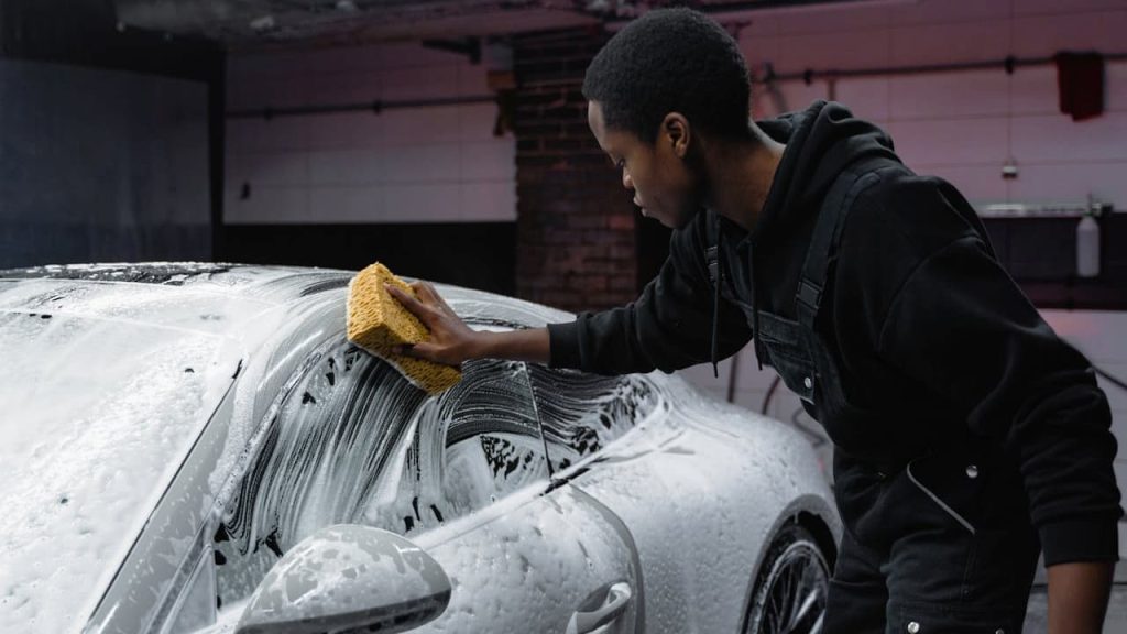 Person in black hoodie washing white car covered in soap suds using yellow microfiber cloth in garage setting