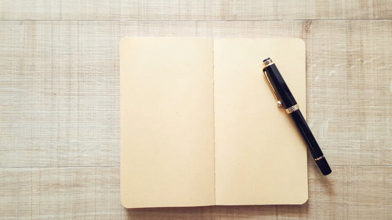 An open blank notebook with cream-colored pages lies next to a black fountain pen on light wooden surface