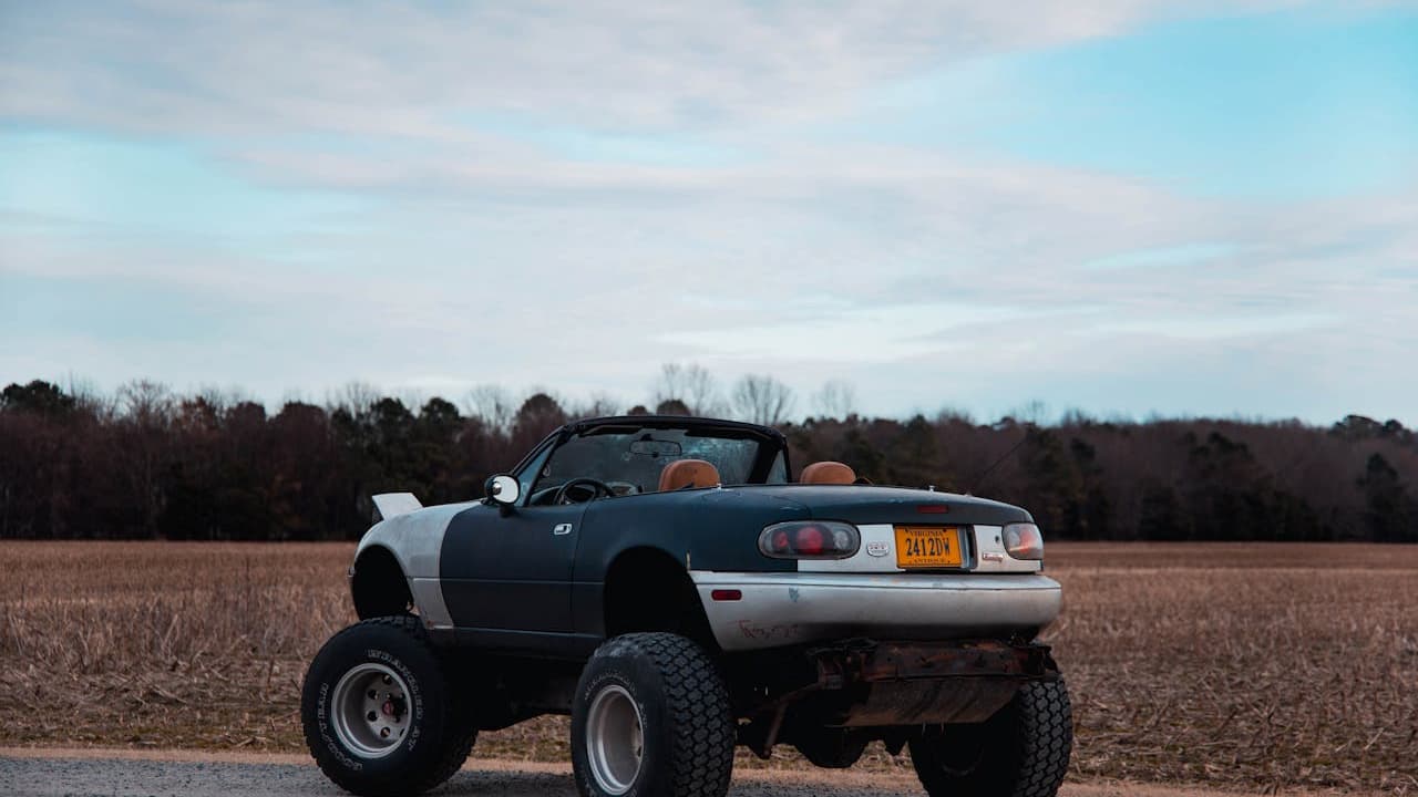 Modified convertible sports car with extremely large oversized wheels and tires in rural field setting under cloudy sky