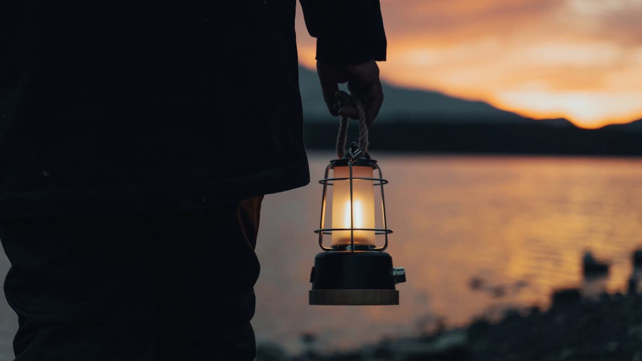 Person holding illuminated camping lantern with metal cage guard against sunset lake backdrop with mountains in silhouette