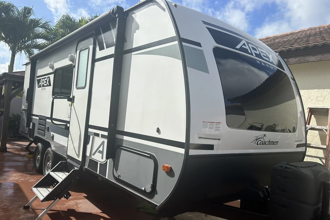 White and gray Coachmen Apex Nano travel trailer with entry steps parked on paved driveway beside house