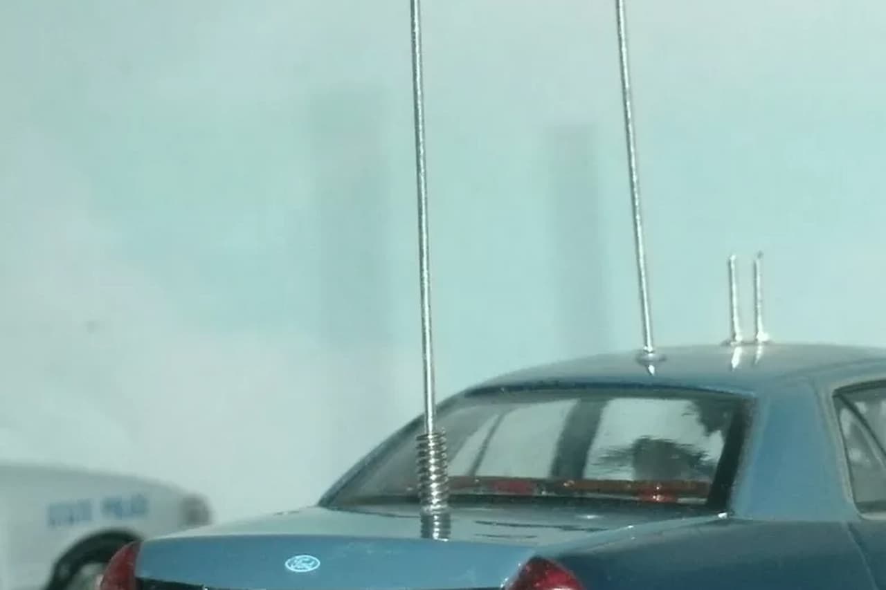 Blue Unmarked Ford police vehicle with multiple tall radio antennas mounted on roof, parked with other patrol cars nearby
