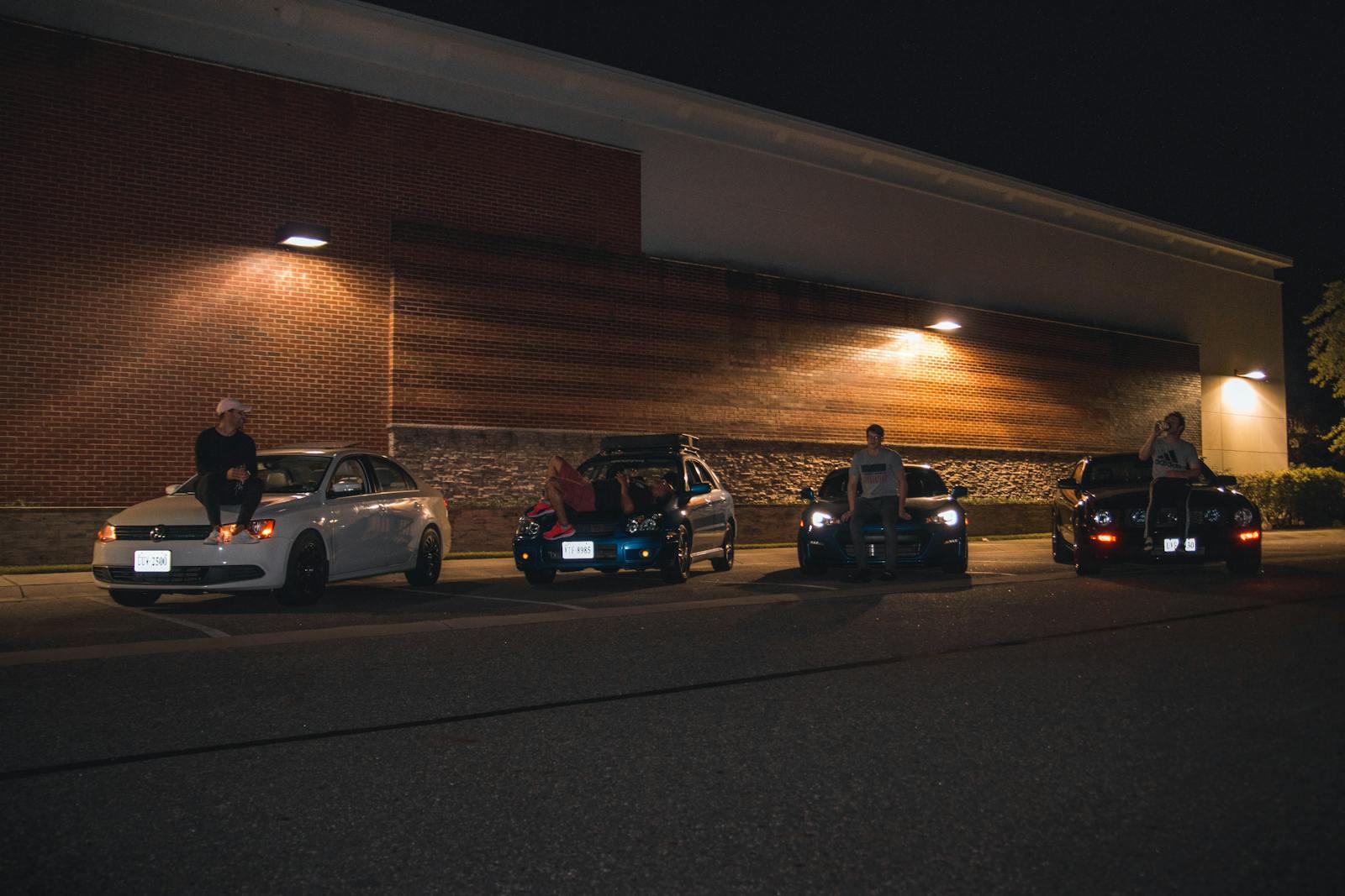 A group of friends sitting on cars in a dimly lit parking lot, enjoying a night out.