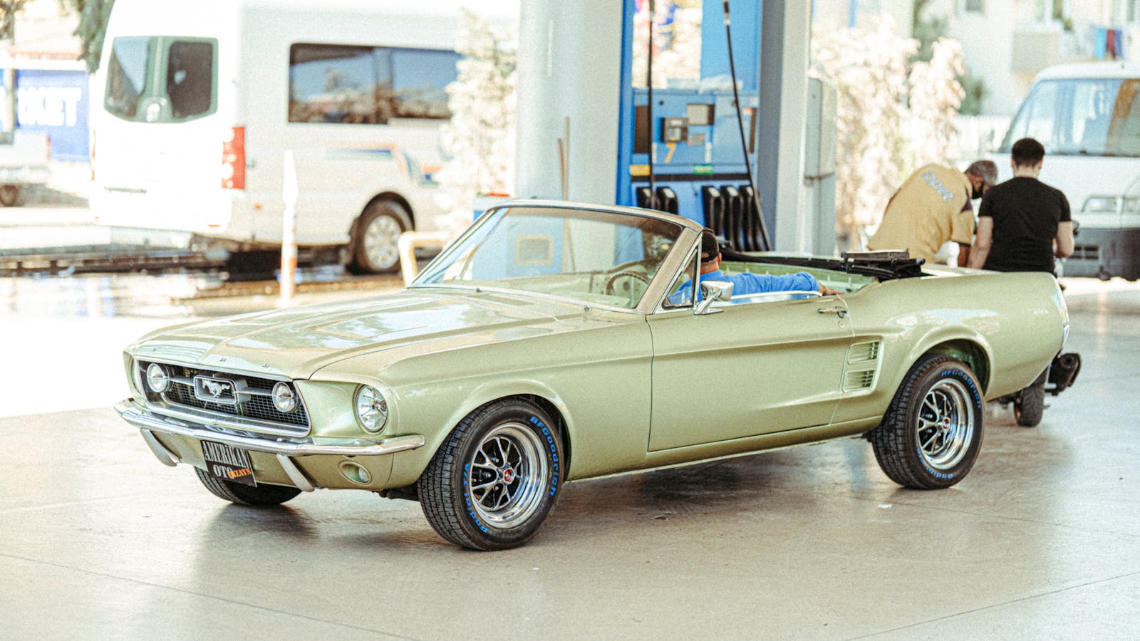 Classic Ford Mustang convertible parked at a Turkish gas station with people around.