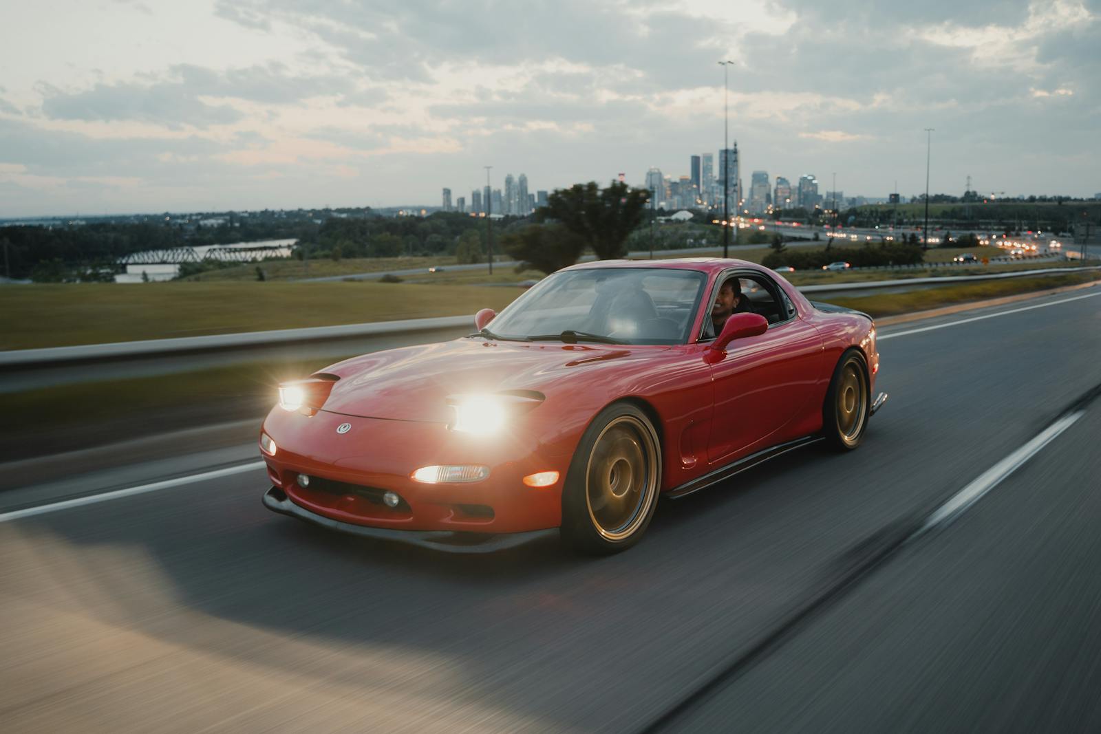 A red Mazda RX-7 sports car driving on a highway with Calgary skyline in the background.