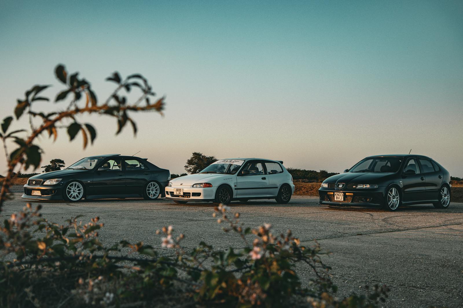 Three modified sports cars parked outdoors at sunset, showcasing tuning culture.