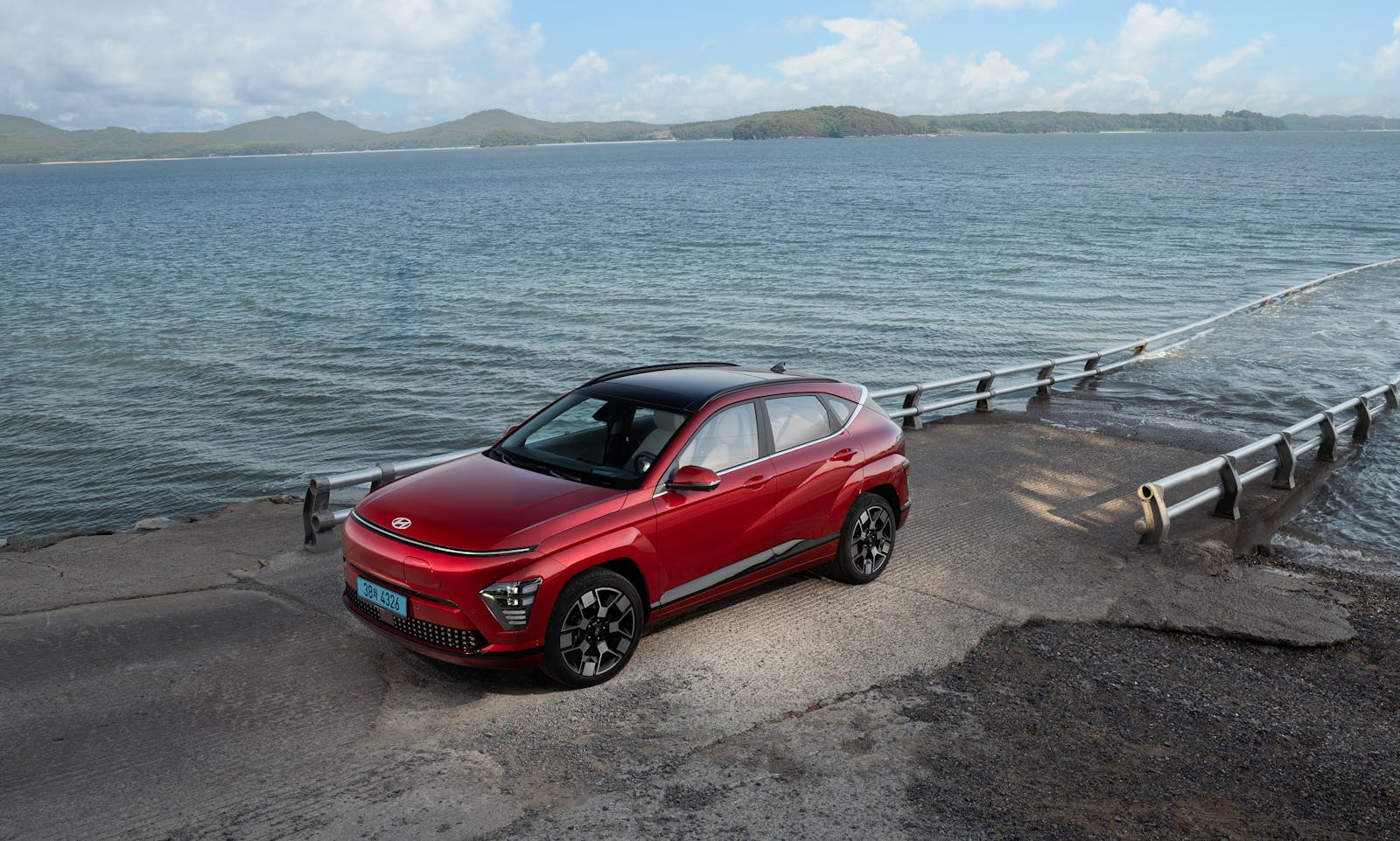 A modern red electric car parked beside a serene seaside road with calm waters and hills in the background.