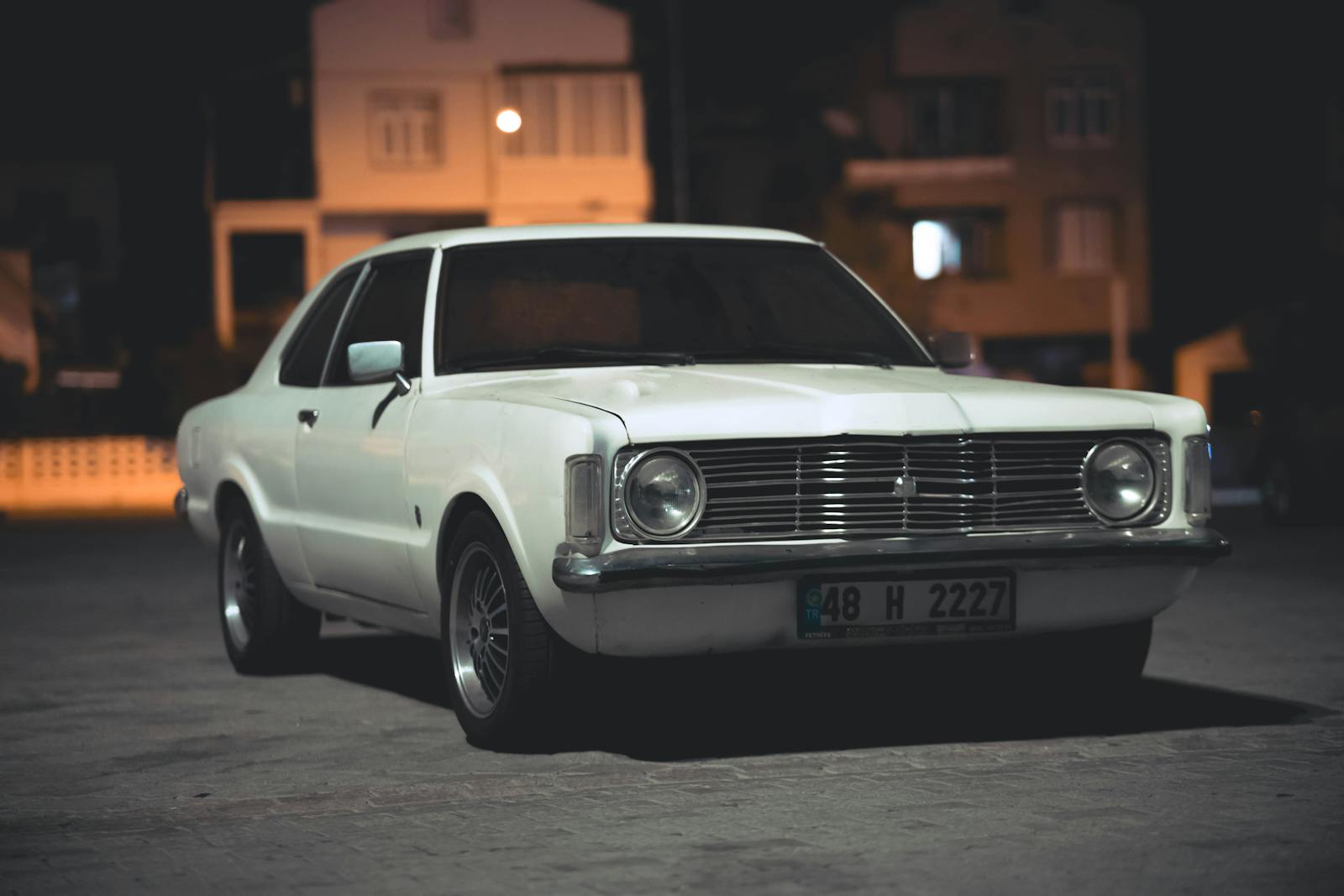 Vintage white Ford Taunus car parked on a dimly lit urban street at night, capturing classic automotive style.