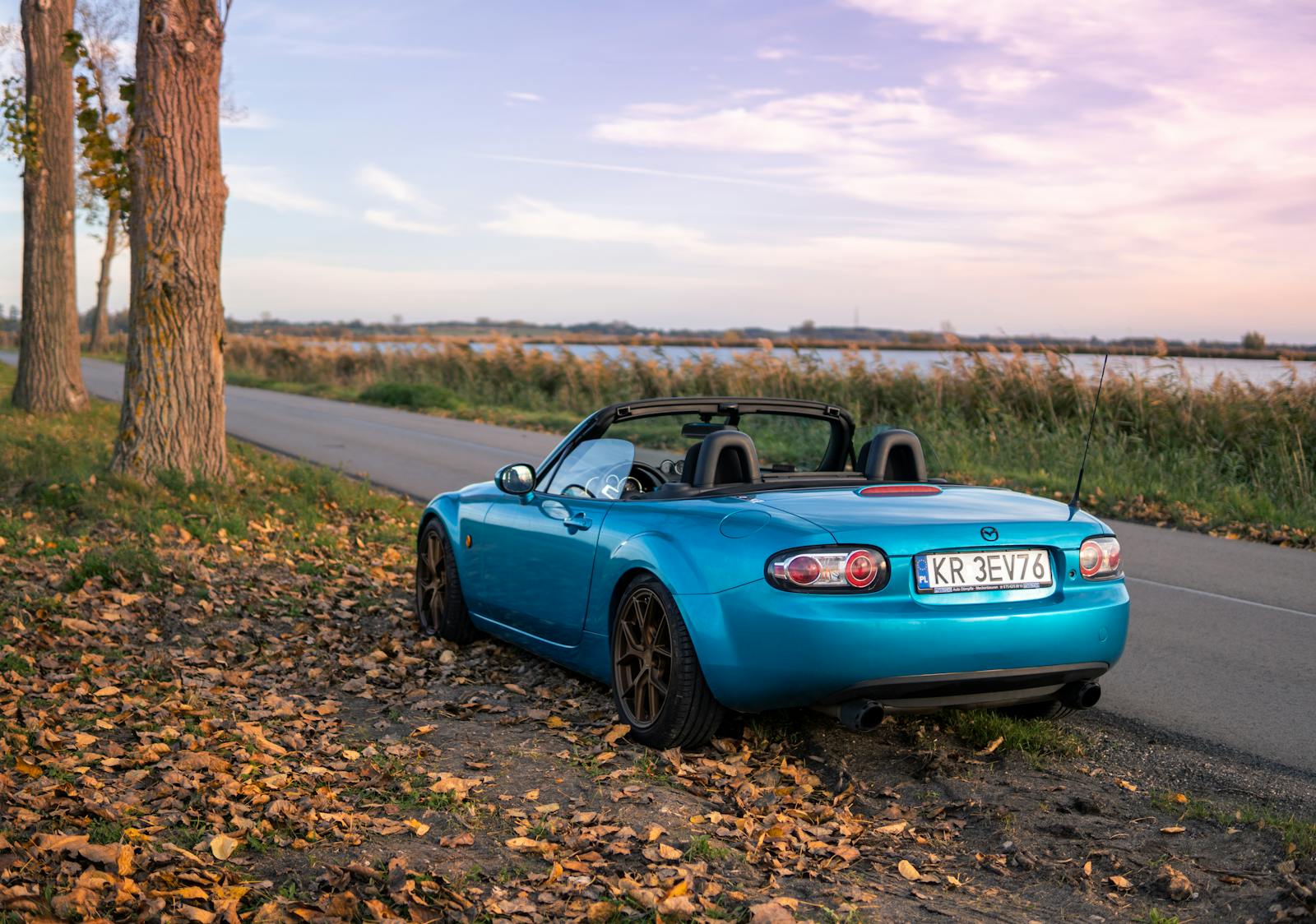 Blue Mazda MX-5 convertible parked by a scenic road in Szczerbaków, Poland during sunset.