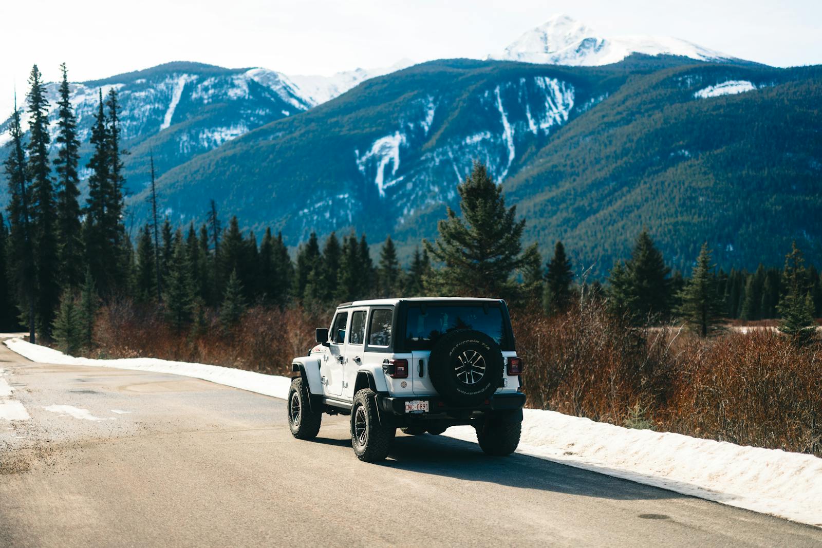 White Jeep on a mountain road in Jasper National Park, Alberta, surrounded by pine trees and snow-capped peaks.