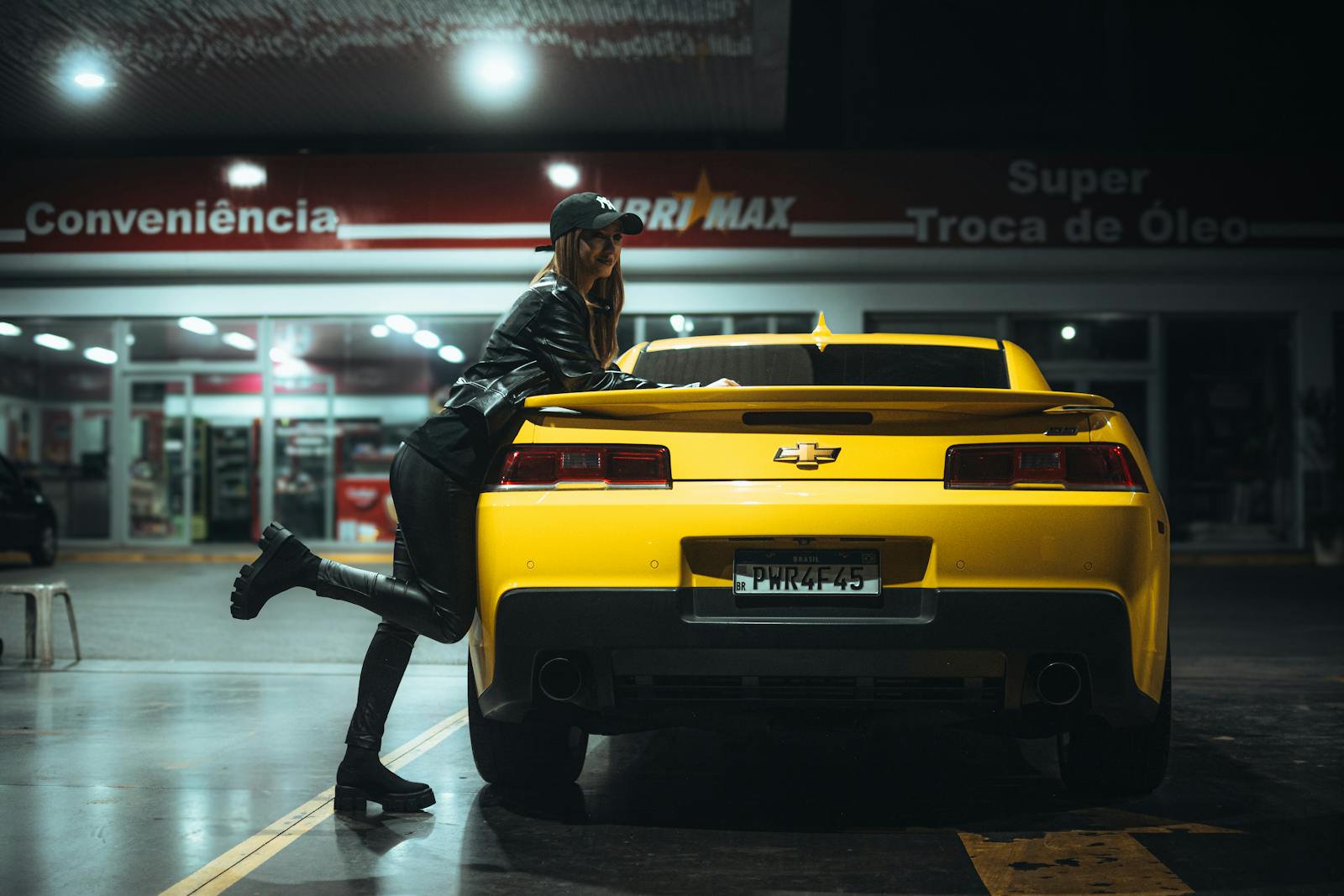 Woman in leather poses with yellow sports car at night gas station.