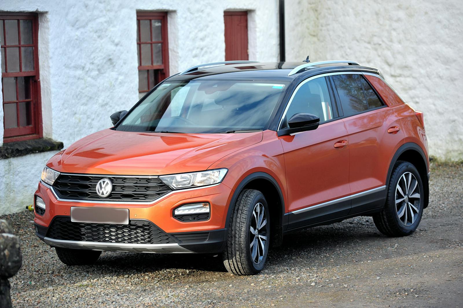 Orange Volkswagen SUV parked on gravel near rustic white building with red windows.